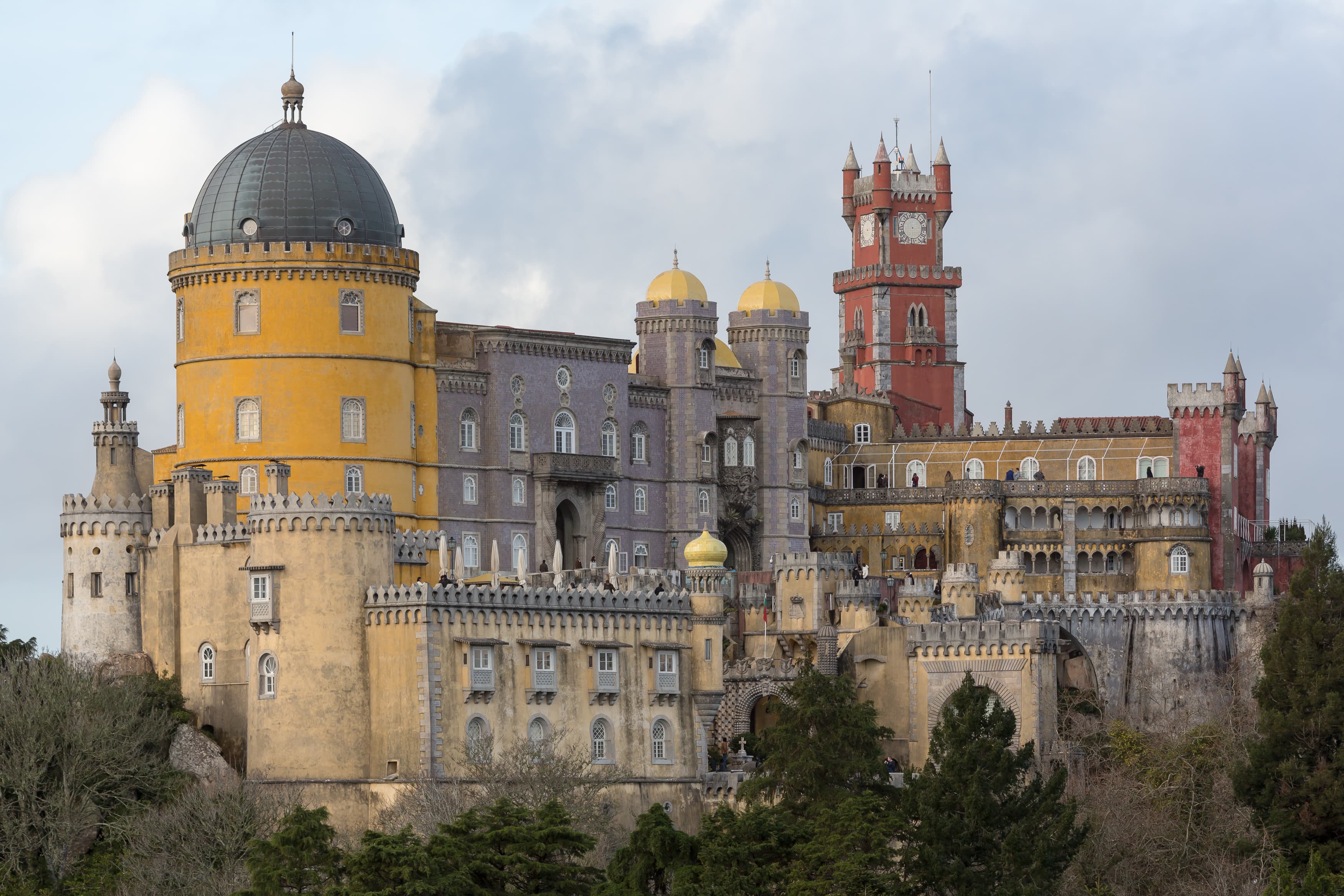 Fachada del Palacio de la Pena con torres y muros pintados en rojo y amarillo, coronando una colina boscosa de Sintra.
